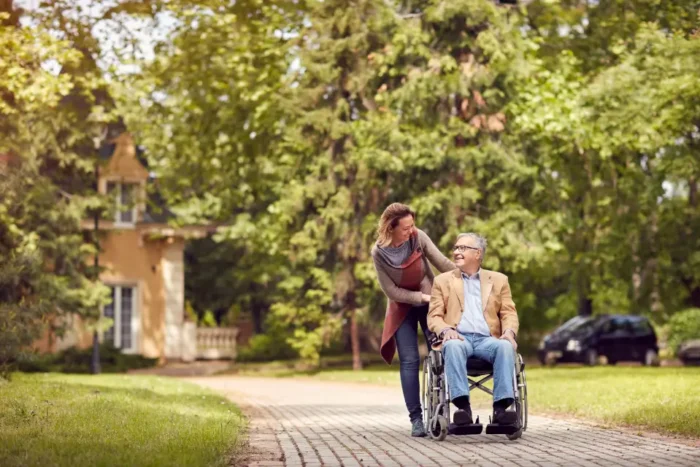 A man sits in a wheelchair outside being pushed by a woman as they have a conversation.