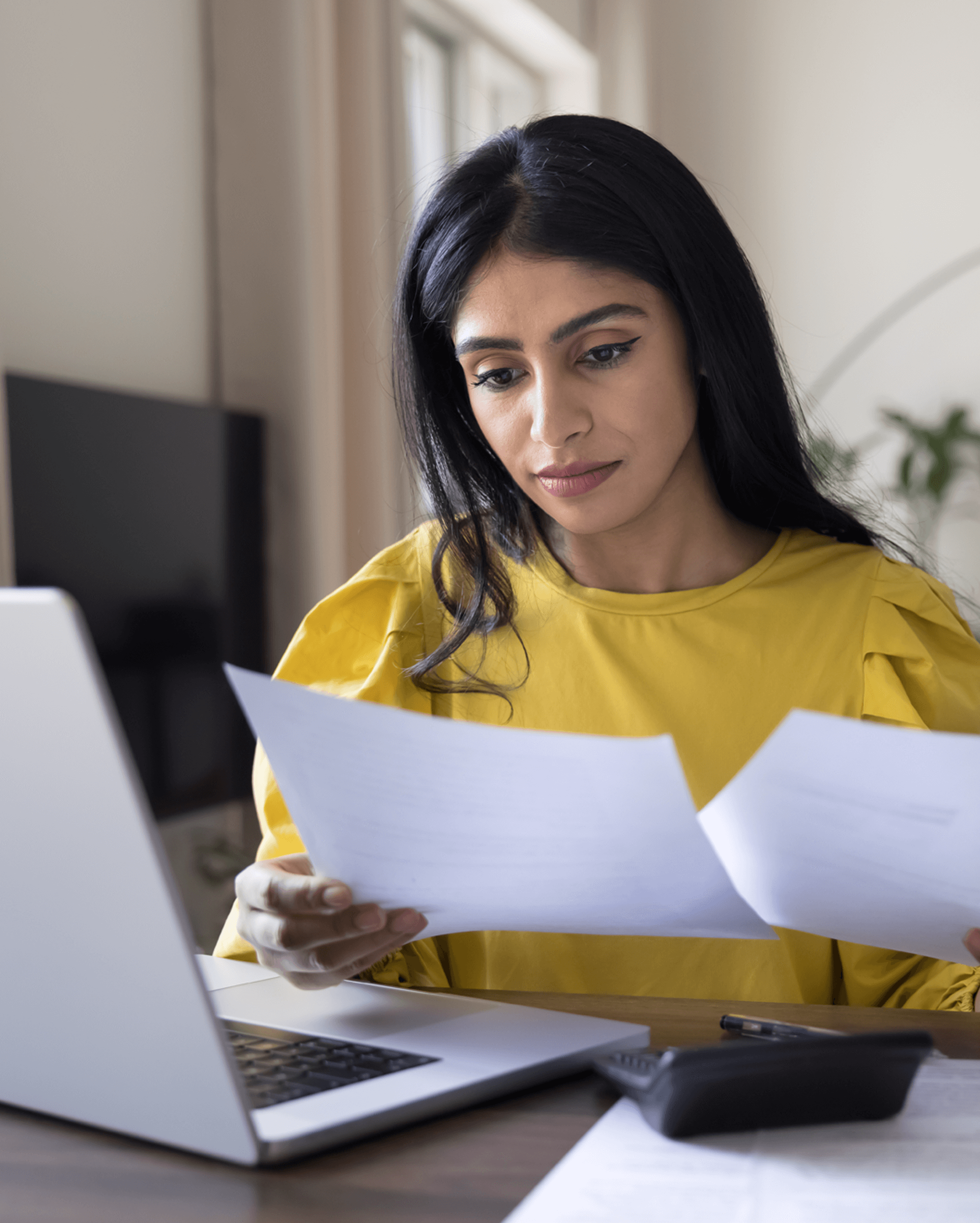 A woman sits at a table in her living room with a laptop in front of her whilst holding a piece of paper in each hand.