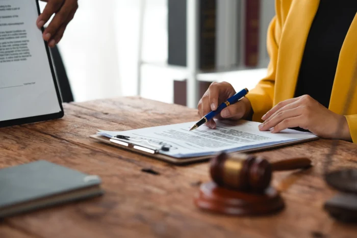 A woman in a yellow blazer sits reviewing a document preparing to sign it with a pen.
