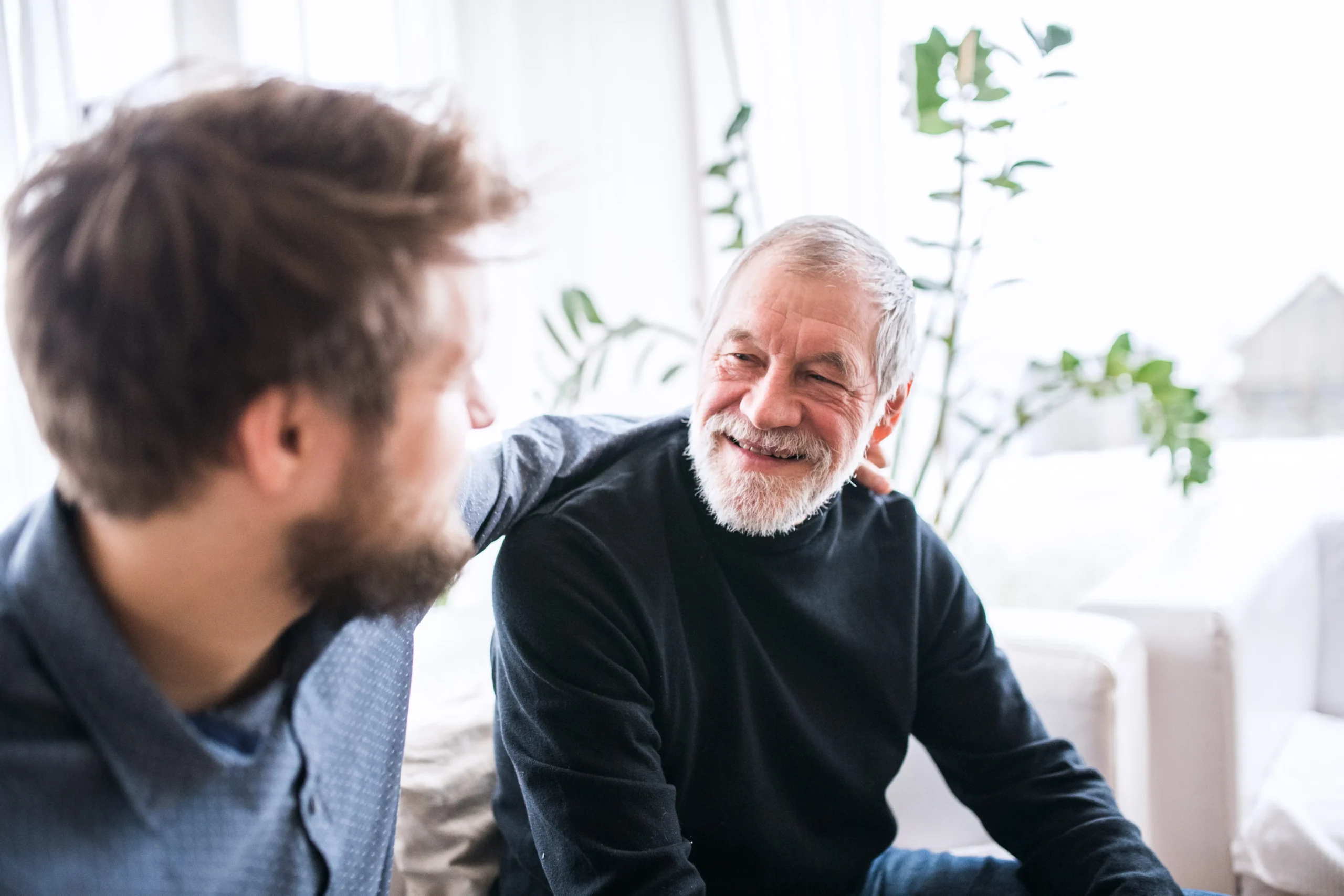 A man puts his arm around an elderly gentleman as they have a conversation on a sofa.