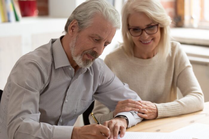 An elderly couple sits at a table filling out a paper form with a pen.