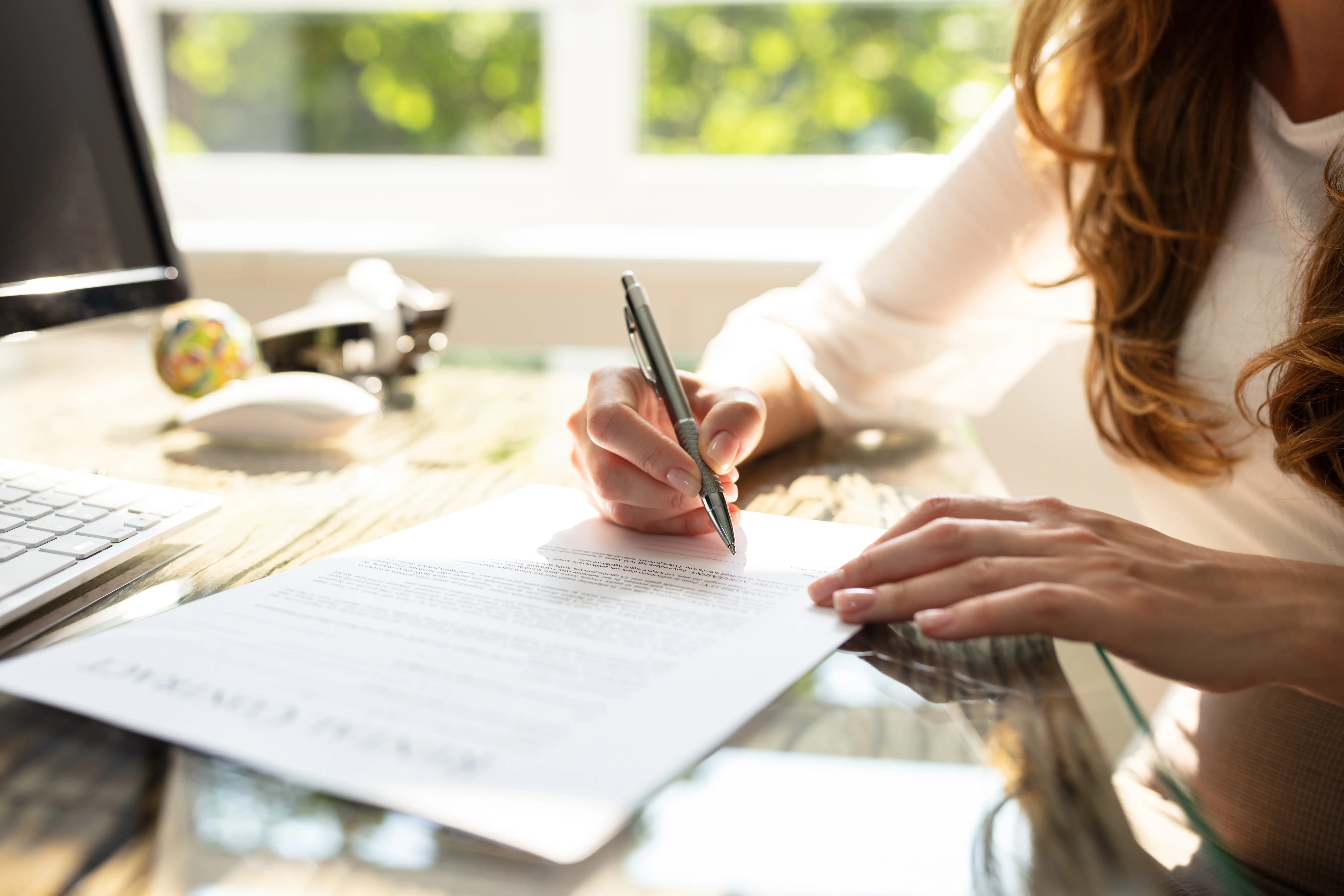 A woman sits as a glass table signing a document with a pen