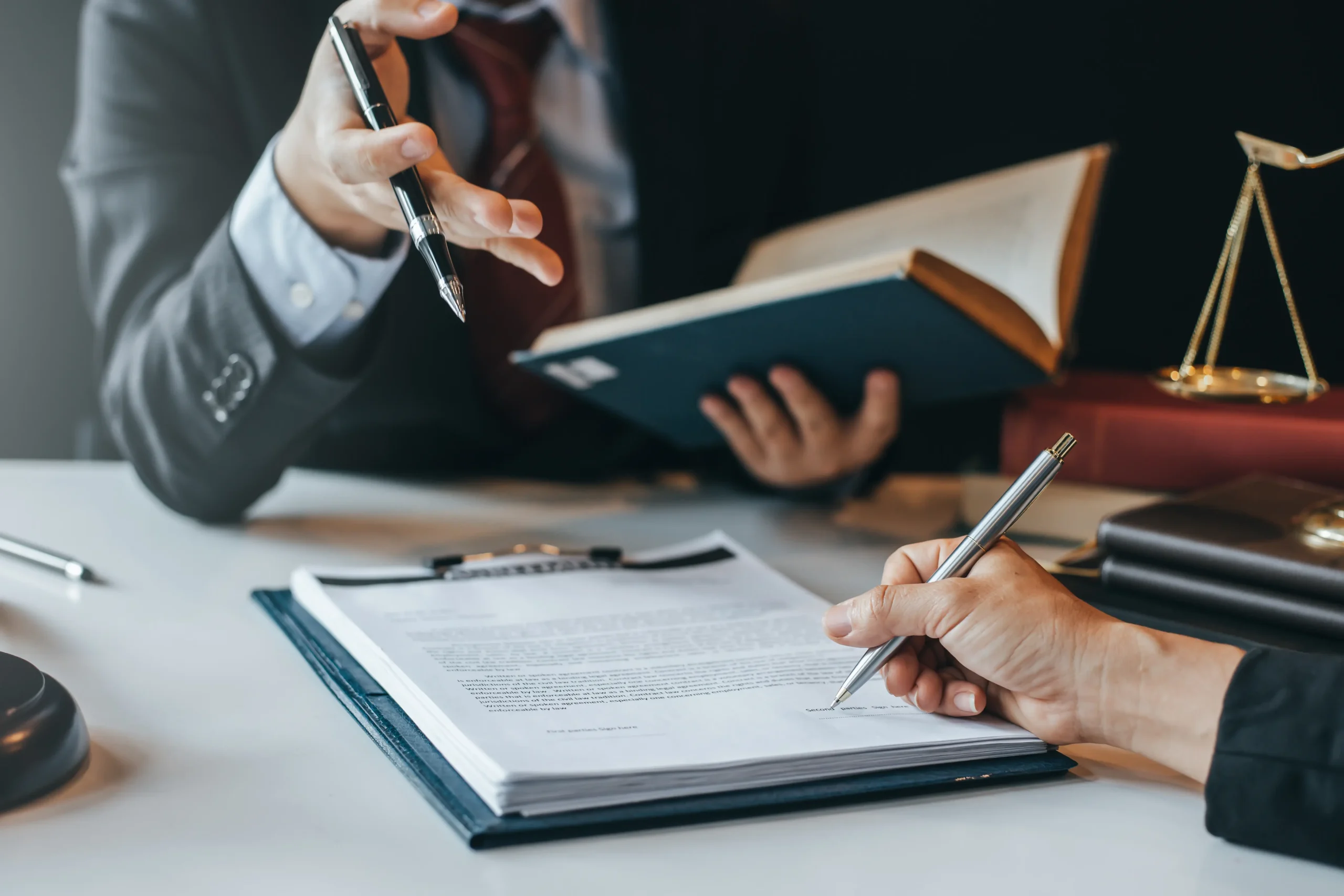 A person is signing a letter with a pen whilst another person in the background holds an open book and has a conversation.