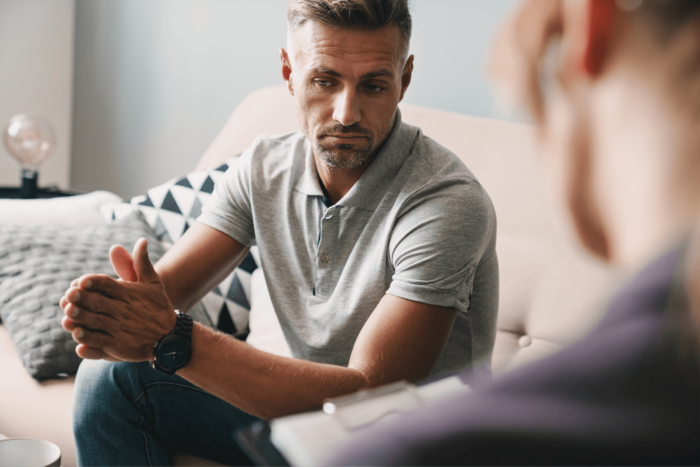 A man sit on a sofa with his hands pressed together whilst talking to someone out of focus in the foreground.