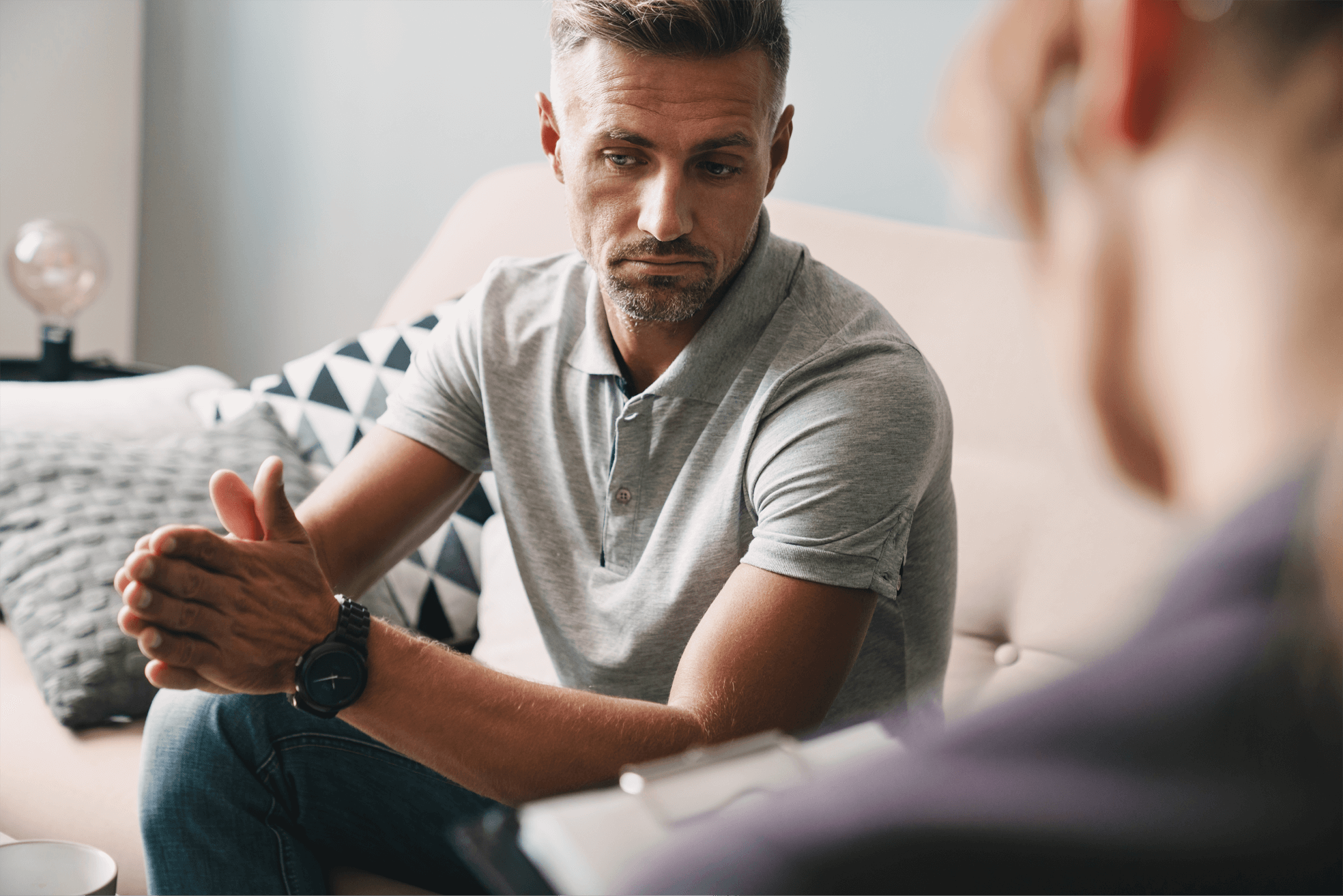 A man sit on a sofa with his hands pressed together whilst talking to someone out of focus in the foreground.