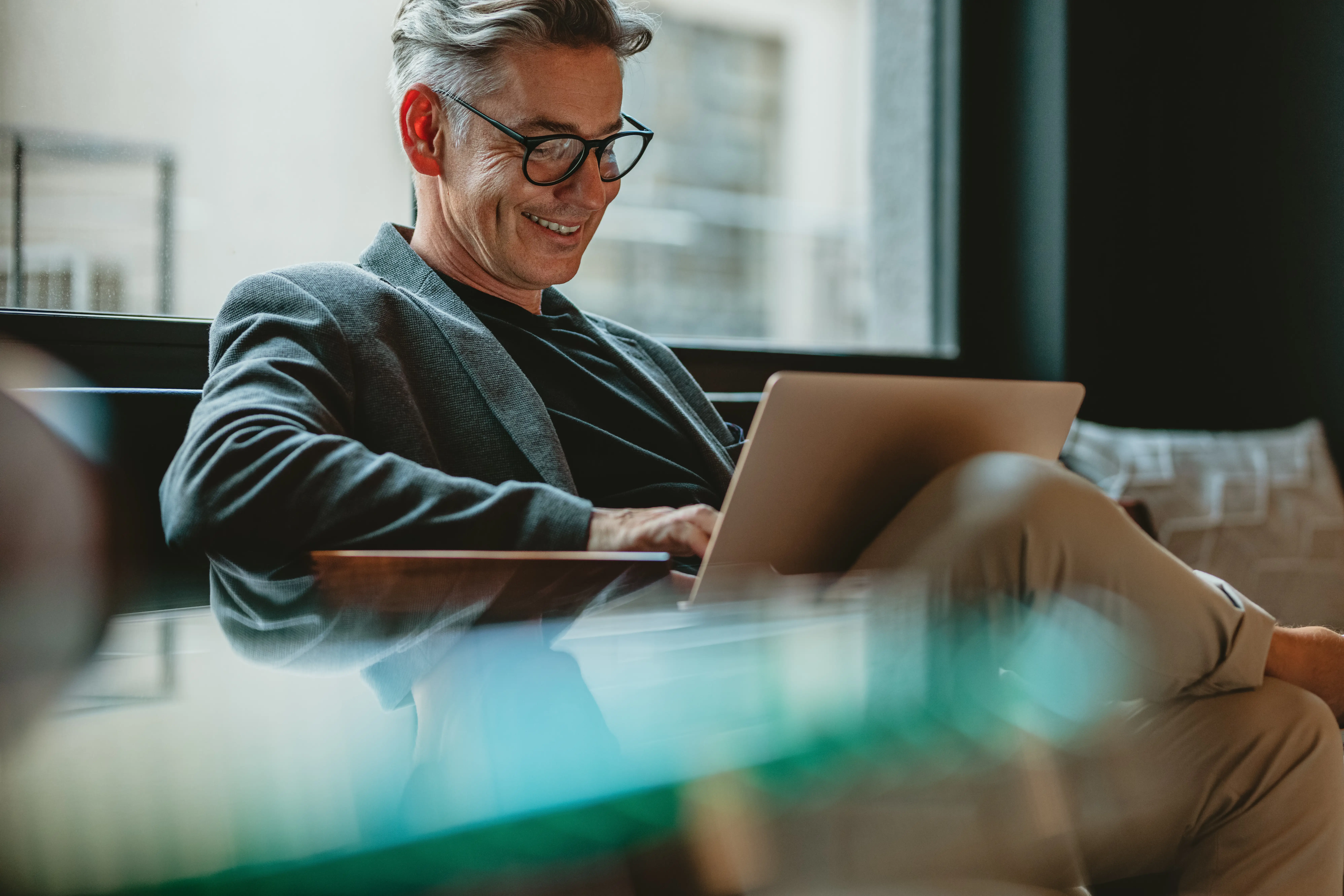 A middle aged man with greying hair sits casually with a laptop in-front of a window.