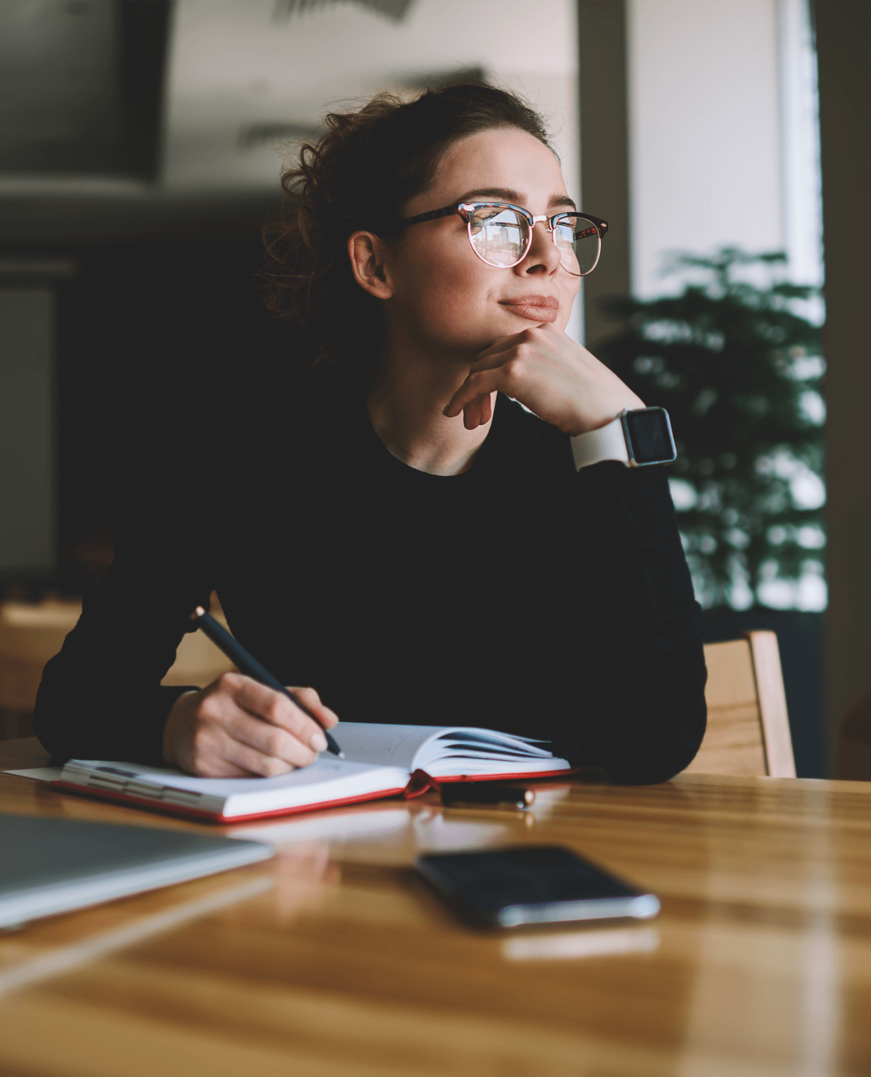 A woman pauses writing in a notebook and looks into the distance.