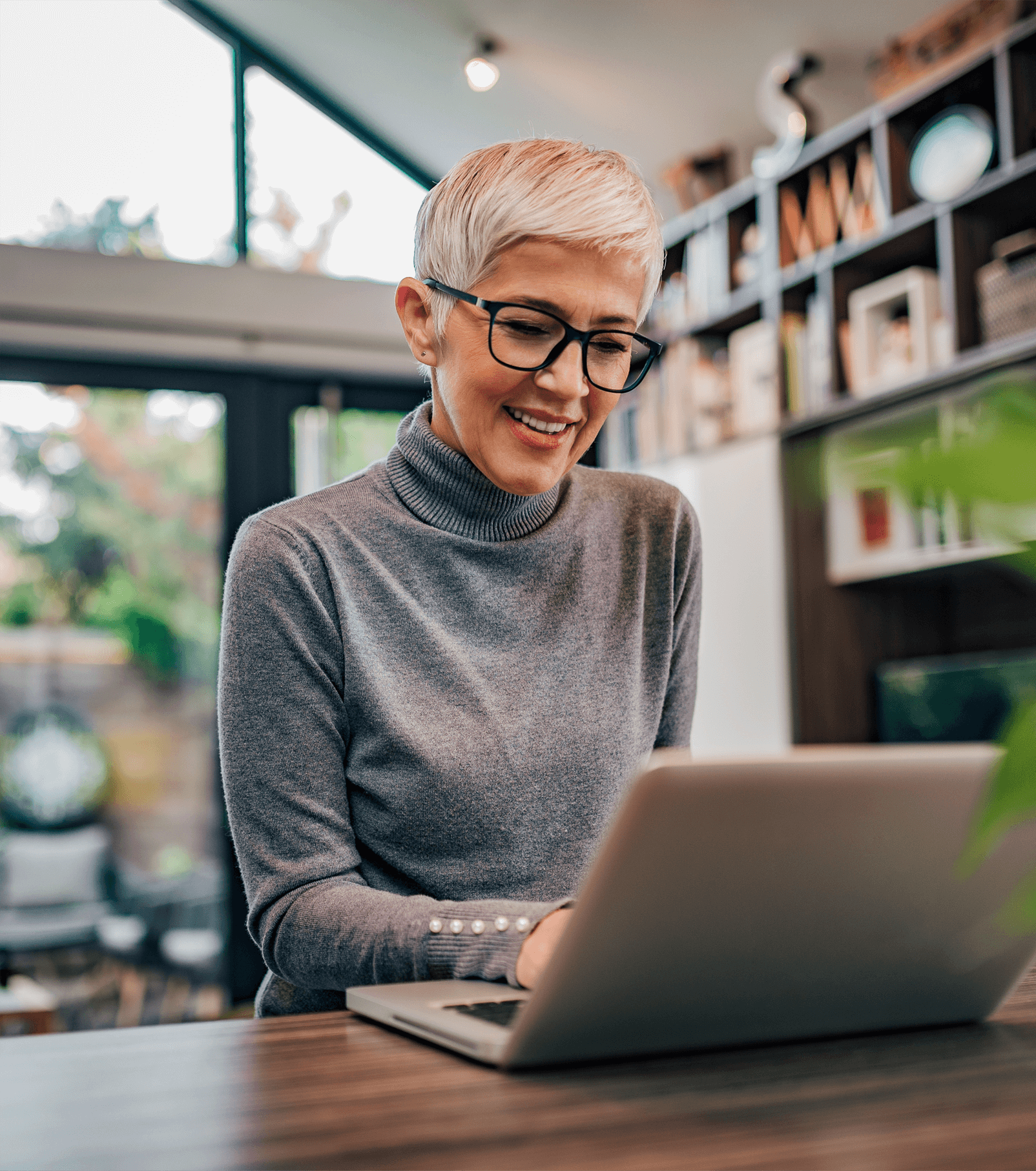 A woman with short greying hair wearing glasses sits at a laptop in her home.
