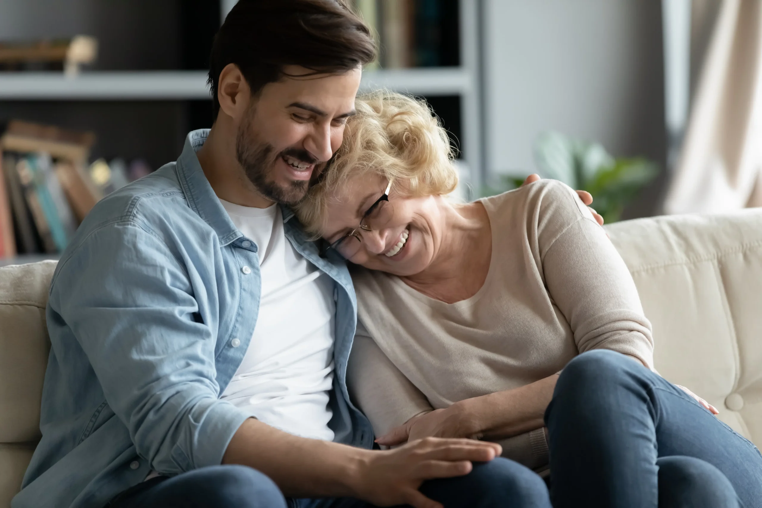 A man sits on the sofa cuddling a middle aged woman as they sit laughing.