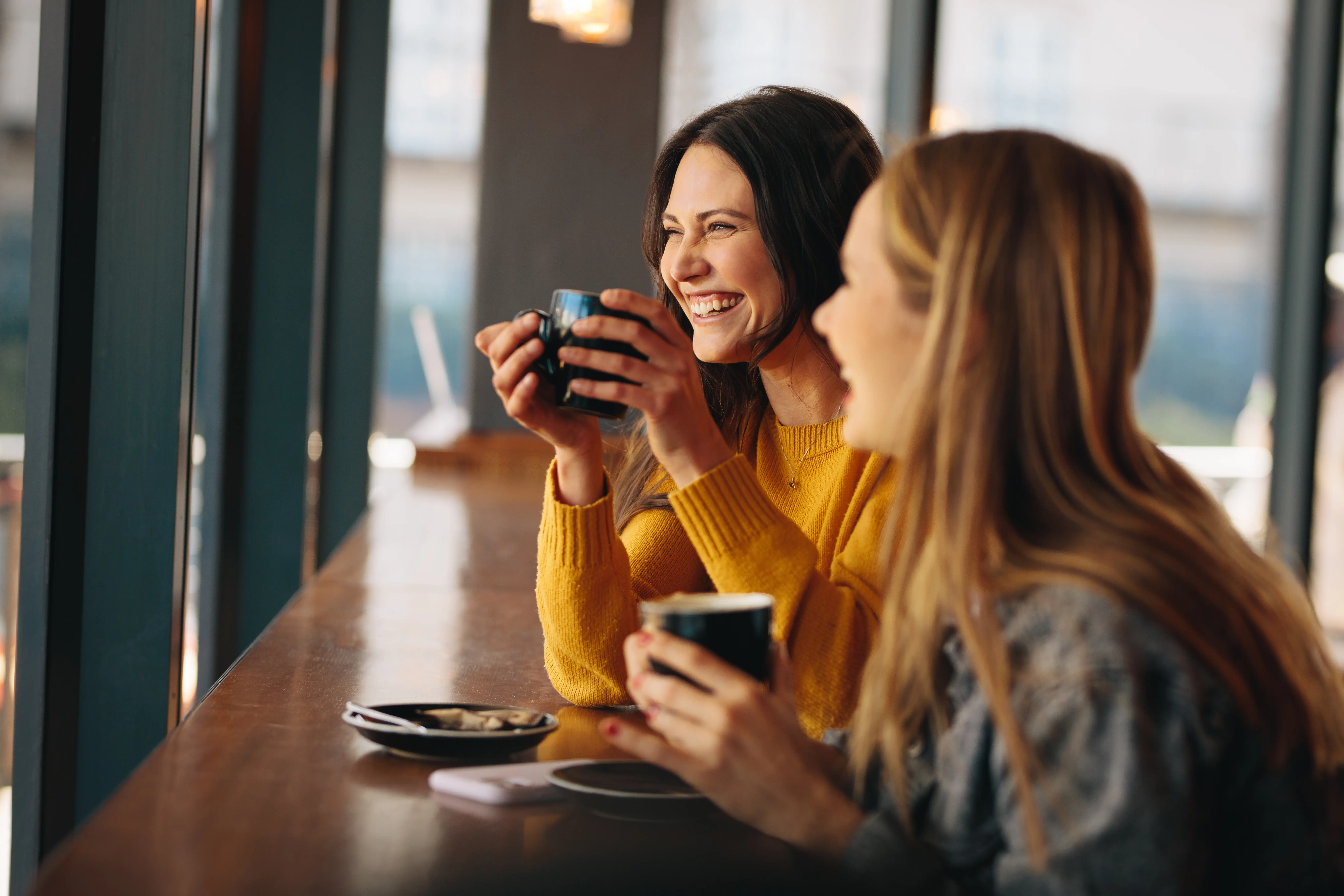 Two women sit laughing over a cup of coffee in a coffee shop.