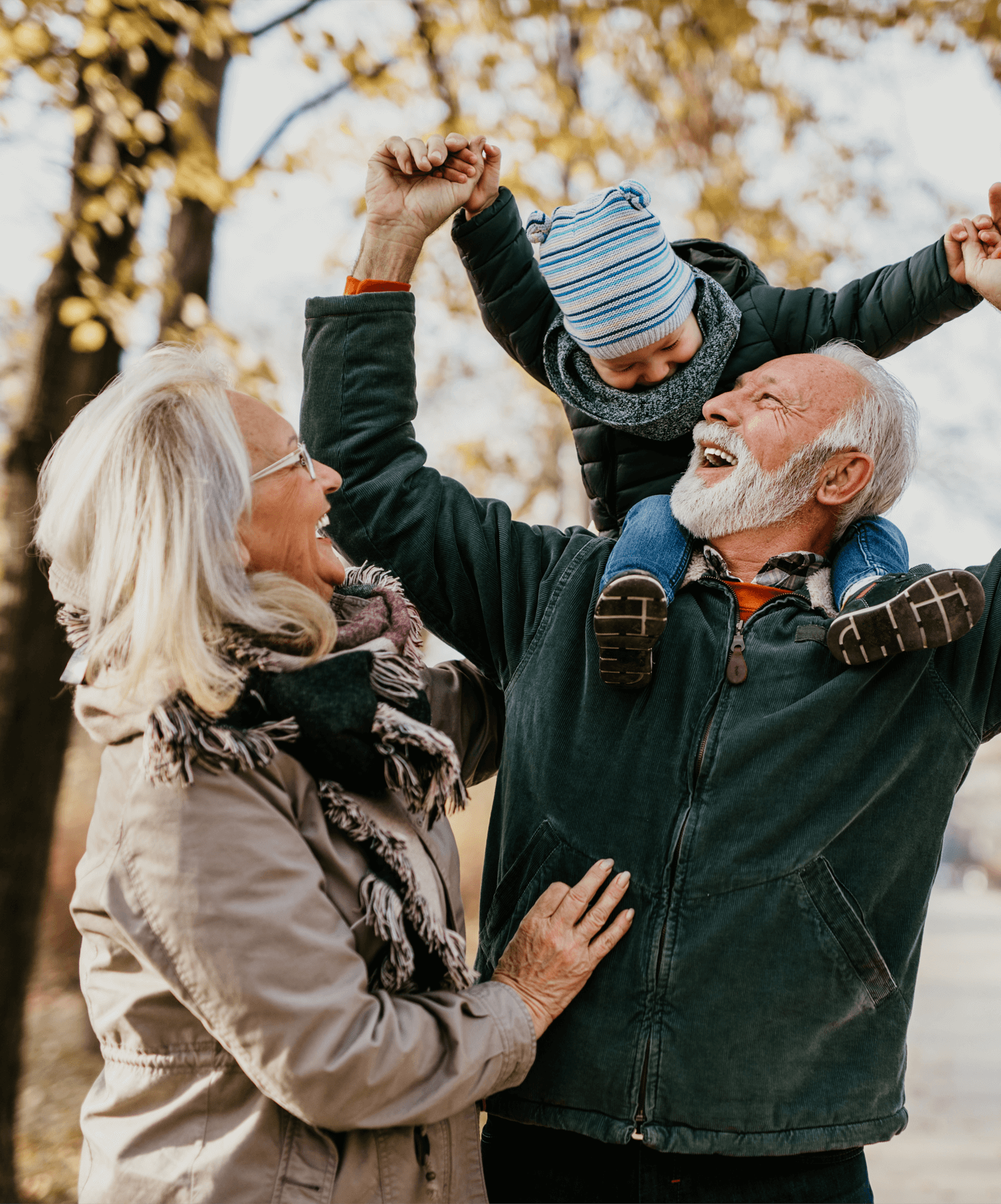A grandfather has his grandson on his shoulders whilst the grandmother stands with them smiling.