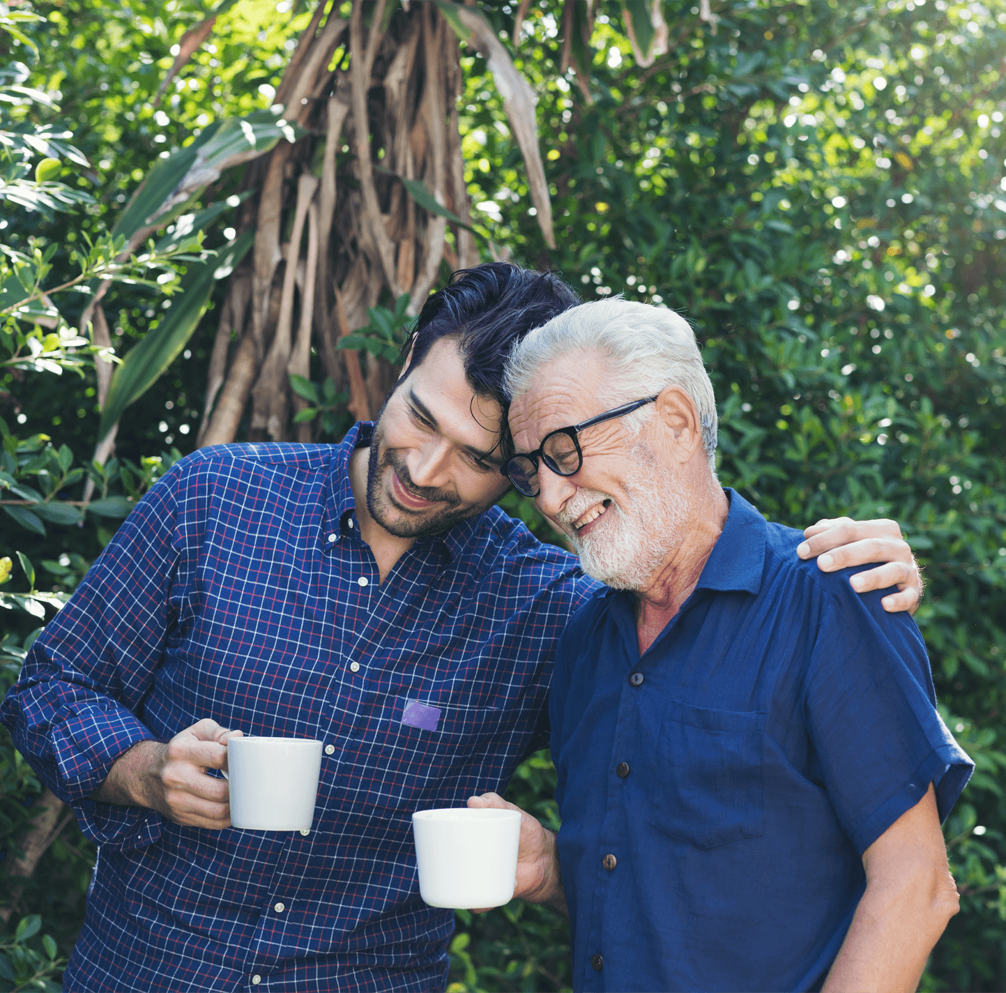 A man and his father stand with their heads together whilst having a coffee.