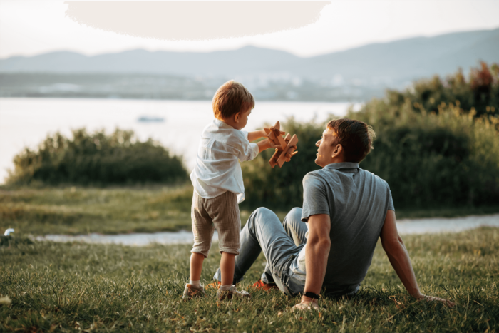 A dad and his son sit looking out over a lake whilst the son plays with his toy wooden airplane.