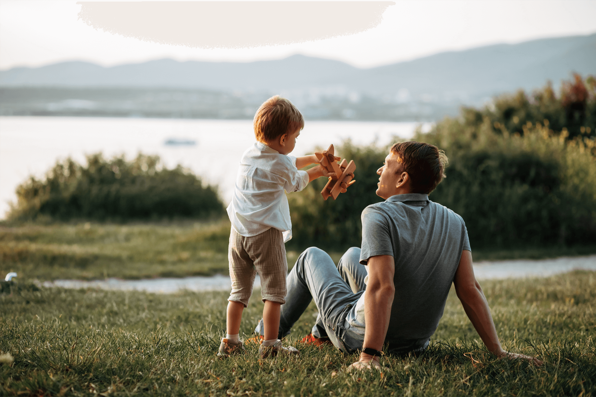 A dad and his son sit looking out over a lake whilst the son plays with his toy wooden airplane.