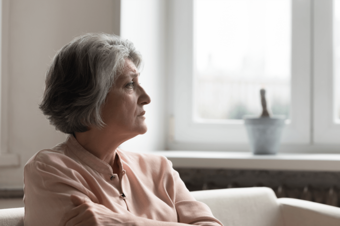 An elderly woman sits alone looking away from the camera out of a window.