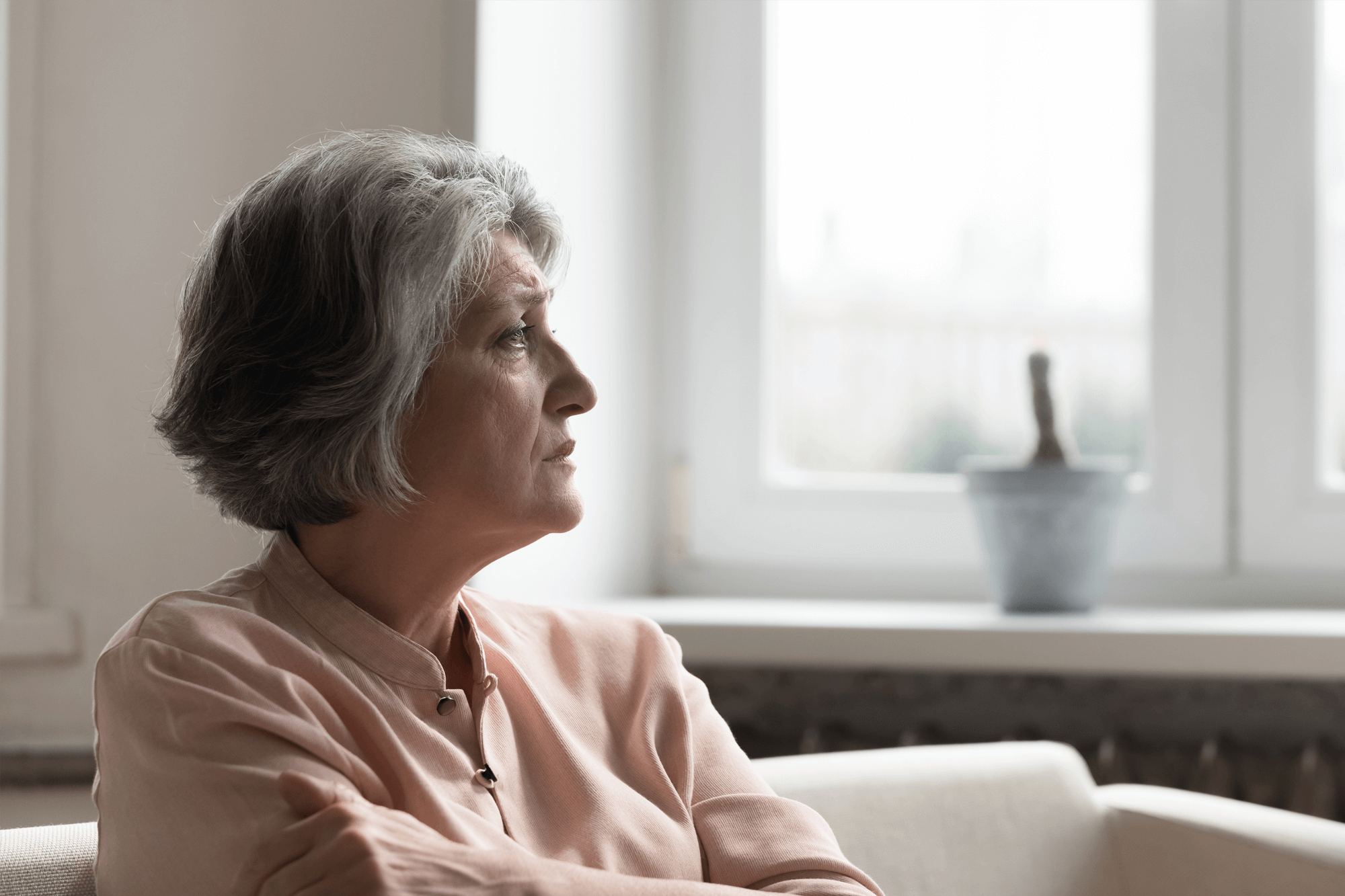 An elderly woman sits alone looking away from the camera out of a window.