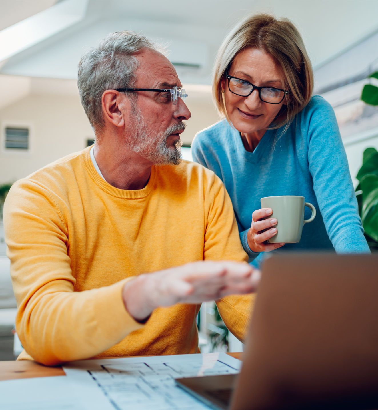 A man sits at a laptop whilst a woman holding a coffee looks a the laptop over his shoulder.