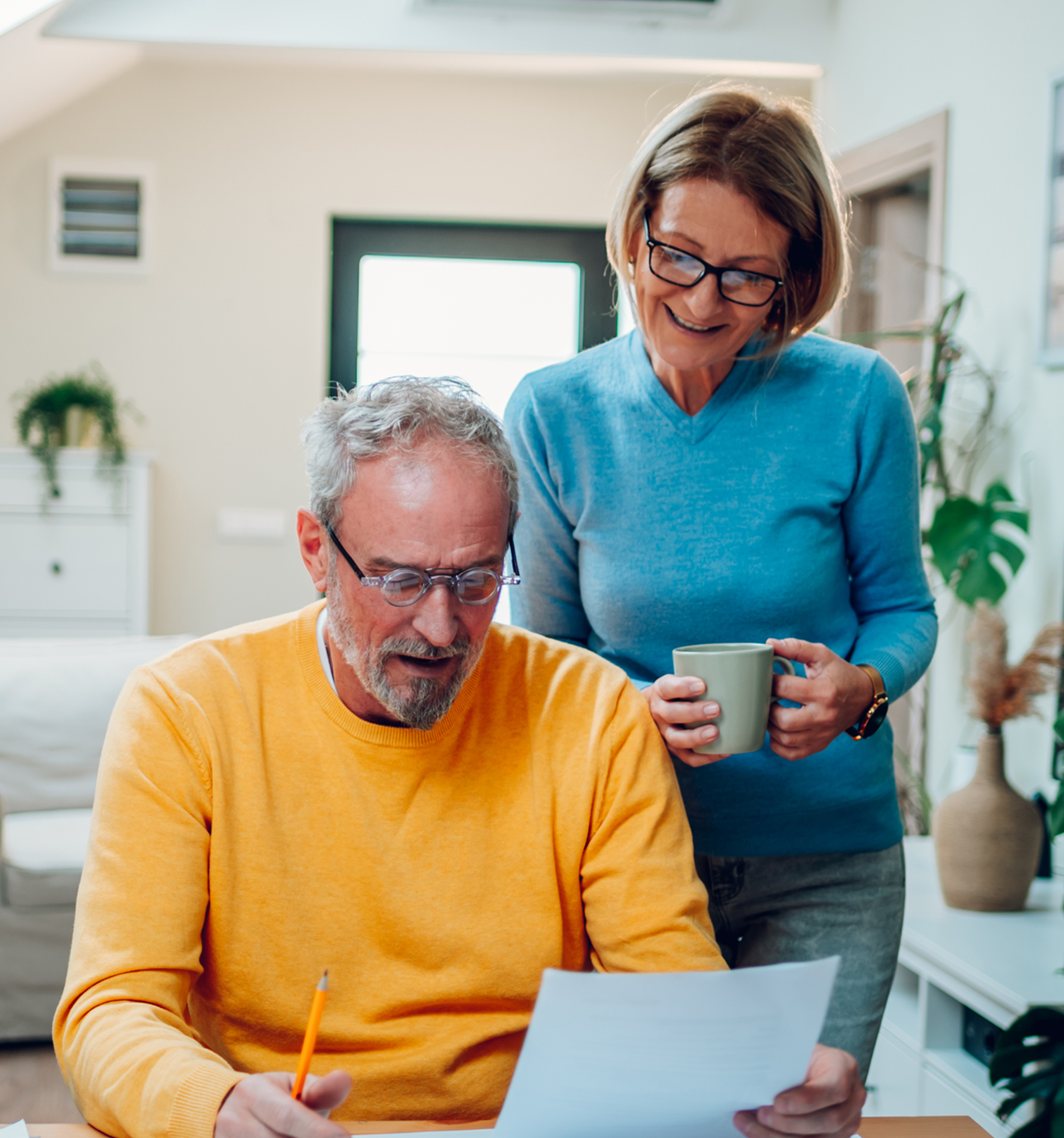A man sits whilst holding a piece of paper and pencil whilst a woman holding a coffee cup stands behind him looking at the papers.
