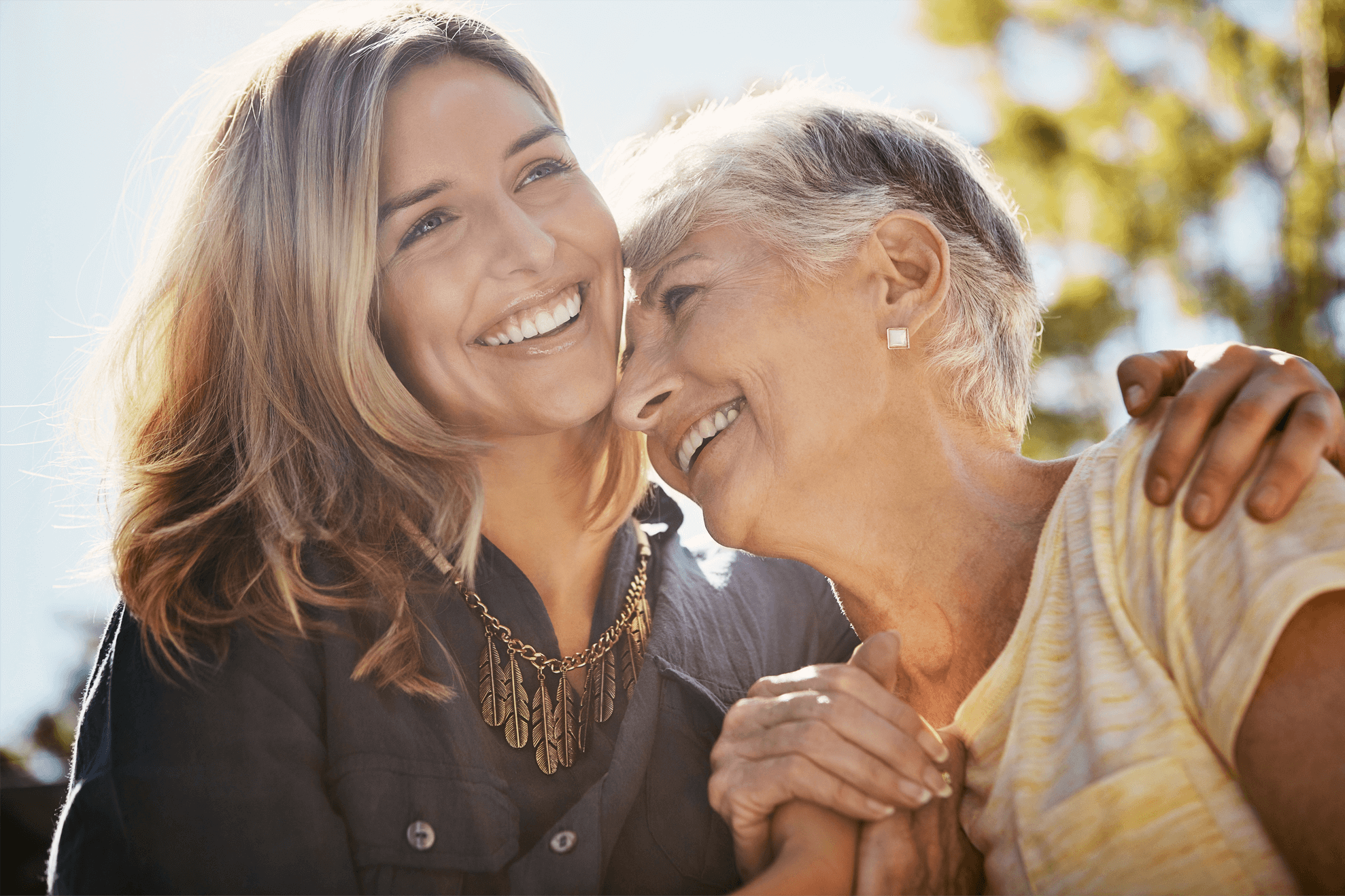 A woman and her mother lean in together whilst smiling and clasping hands.