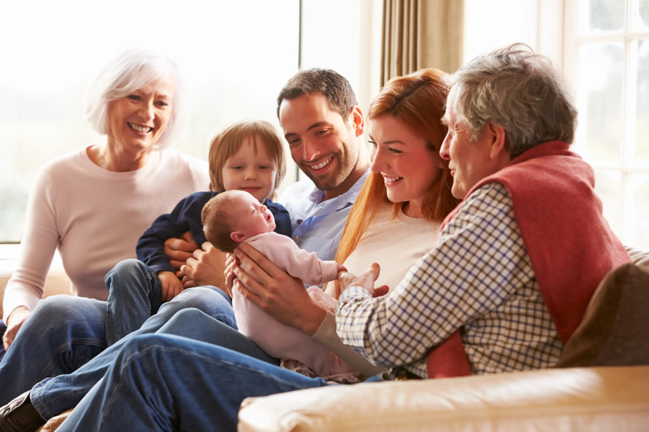 A multi-generational family are seated in a living room greeting a new baby.