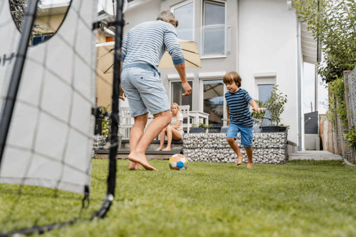A father and son play football in the garden whilst the mum watches from the patio.