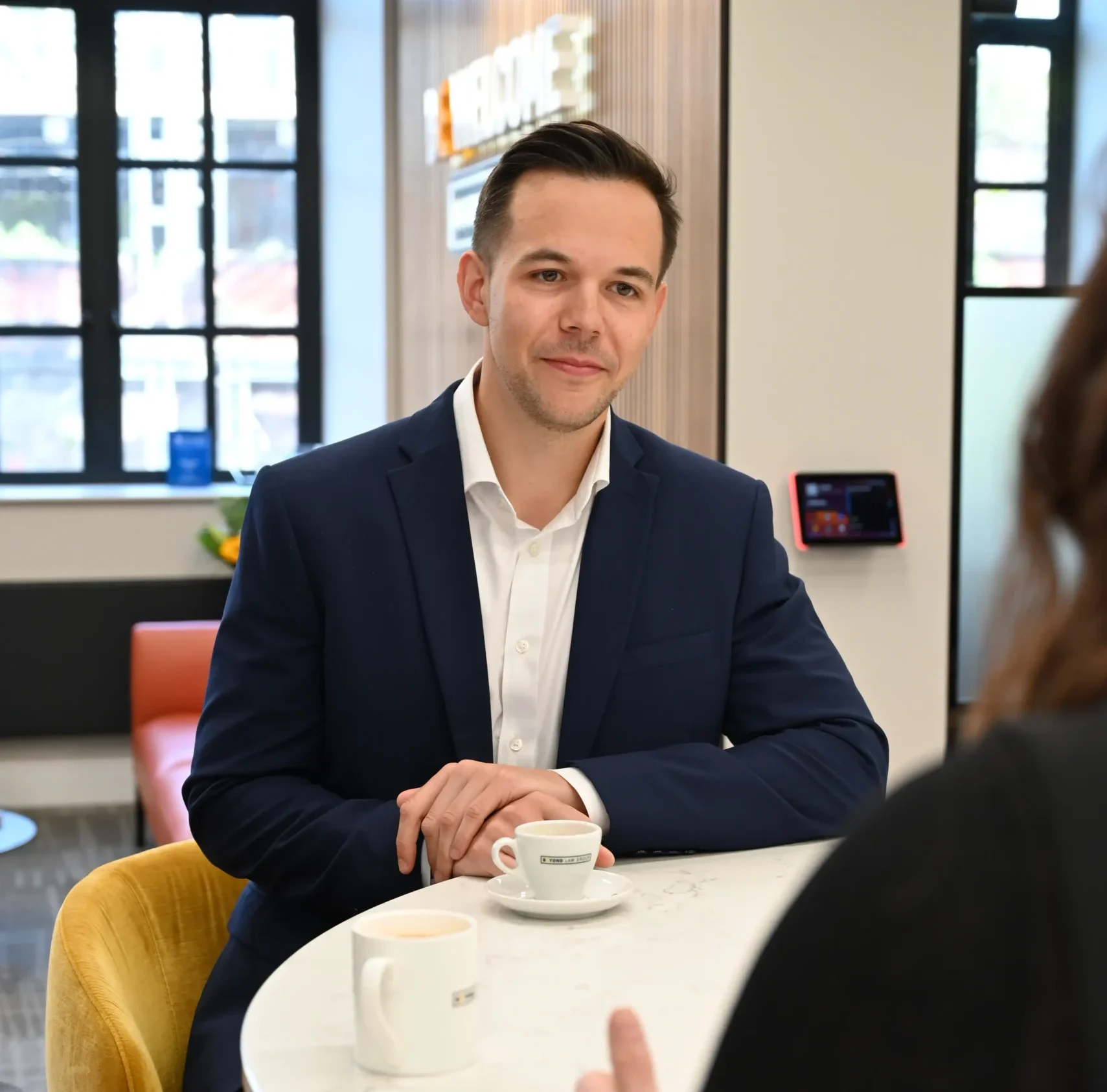 Russell Kaminski sits at table having a conversation with someone out of shot with coffees on the table.